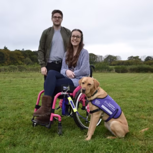 Photo of a man, a smiling woman in a wheelchair, & a service dog, posing for a photo in the middle of a large, grassy field.